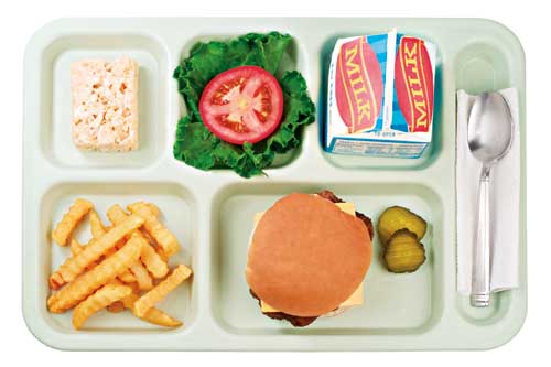 A top-down view of a school lunch tray, featuring milk, a burger with pickles, crinkle-cut fries, a Rice Krispies treat, lettuce and tomato.