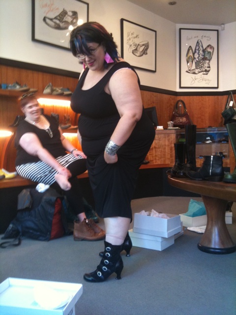 Marianne, in a long black tank dress, tries on Fluevog boots, with are black with many little buckles.